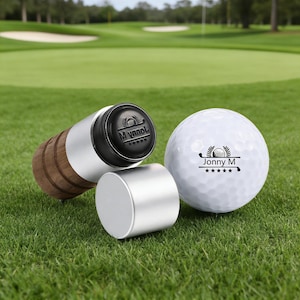 Peut inclure: Un ensemble de marqueurs de balle de golf et une balle de golf sur de l'herbe verte. La balle de golf est blanche avec le texte "Jonny M" et un logo. L'ensemble de marqueurs comprend un marqueur argenté et bois avec le texte "M Vynol".