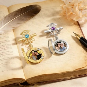 May include: Two graduation-themed locket pendants. One is gold-toned with a blue gemstone, the other silver-toned with a purple gemstone. Both feature a graduation cap, the year "2026", and a space for a photo. The lockets are open, revealing photos.