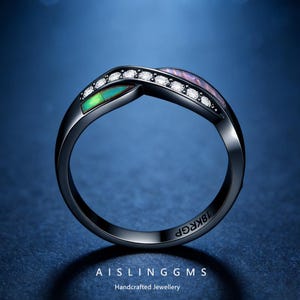 May include: Black ring with a twisted design, featuring a row of small white stones and two sections of colourful, iridescent material. The ring has the text "18K RGP" and "AISLING GMS Handcrafted Jewellery" engraved on it.