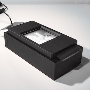 May include: A black rectangular device, possibly a digital photo frame, with a screen sits on a white surface. A black cord and plug extend from the device. A white clock with black numbers is in the background.