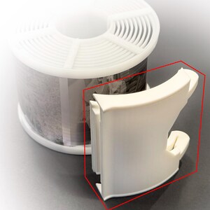May include: A white plastic 3D printed component with a curved, bracket-like shape featuring a vertical slot, positioned next to a white plastic spool containing a roll of black and white photographic film with visible imagery.