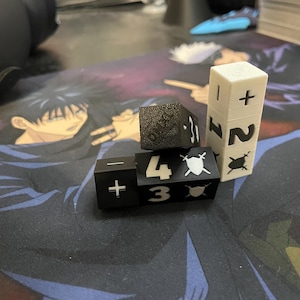 May include: A collection of dice, including black and white cubes with symbols and numbers. The dice feature plus and minus signs, numbers, and sword icons. One die is white with numbers and symbols.
