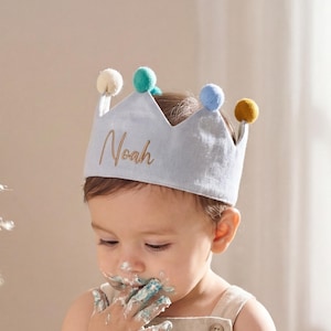 Può includere: Un bambino con una corona azzurra con il nome "Noah" in lettere dorate, che mangia una torta. La corona ha pompon colorati. La torta è su un supporto di legno. Il bambino indossa un pagliaccetto beige.