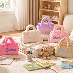 May include: A selection of soft, pastel-coloured handbags with embroidered names: Daisy, Amelia, Bella, Baby, and Chloe. Each bag has a top handle and a shoulder strap. Children's books, crayons, and a teddy bear are also present.