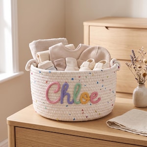May include: A round, off-white rope basket featuring colourful confetti-like flecks and the name Chloe embroidered in pink, purple, green, and yellow yarn. The basket is filled with various neutral-toned baby items, including a blanket, hat, and booties.