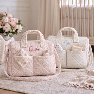 May include: Two quilted fabric nappy bags with top handles and shoulder straps. The pink bag features the embroidered name Olivia, while the cream bag features the name Ava. Both bags have two front pockets containing small cloth items.