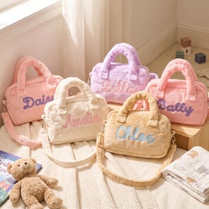 May include: Five pastel-coloured plush handbags with embroidered names: "Daisy", "Amelia", "Hello", "Bally", and "Chloe". Each bag has a matching handle and shoulder strap. A children's book, a teddy bear, and a baby blanket are in the foreground.