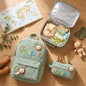 May include: A sage green corduroy backpack and matching pencil case, both personalized with the name Jasper in colorful embroidered letters and a small plush lion patch. The set includes an open insulated lunch bag containing food items.