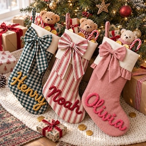Peut inclure: Trois chaussettes de Noël avec des revers crème et de grands nœuds en tissu. La première est à carreaux verts et bleu marine avec l'inscription Henry en laine jaune. La deuxième est rayée rouge et blanc avec l'inscription Noah en laine rouge. La troisième est rose uni avec l'inscription Olivia en laine rose.