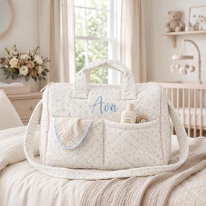 Könnte beinhalten: Eine gesteppte Wickeltasche aus cremefarbenem Stoff mit kleinem blauem Blumenmuster. Die Tasche hat zwei Tragegriffe, einen langen Schulterriemen und zwei Vordertaschen. Der Name Ava ist in blauer Schrift gestickt. Ein Tuch und Lotion sind enthalten.