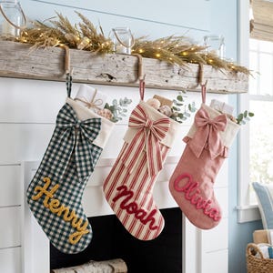 May include: Three Christmas stockings hanging from a wooden mantel. The first is green and white gingham with the name Henry in yellow. The second is red and white striped with the name Noah in red. The third is solid pink with the name Olivia in pink.