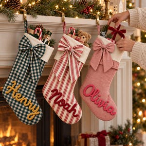 May include: Three Christmas stockings hanging from a mantel. The first is green and white gingham with the name Henry in yellow. The second is red and white striped with the name Noah in red. The third is solid pink with the name Olivia in red.
