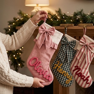 May include: Three personalised Christmas stockings hanging from a wooden mantel. The first is solid dusty pink with a matching bow and the name Olivia. The second is green and white gingham with a bow and the name Henry. The third is red and white striped with a bow and the name Noah.