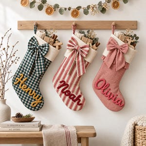 May include: Three Christmas stockings hanging from a wooden rack. The first is green and white gingham with the name Henry in yellow yarn. The second is red and white striped with the name Noah in red yarn. The third is solid pink with the name Olivia in pink yarn.