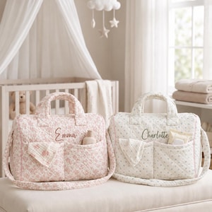 Könnte beinhalten: Zwei gesteppte Wickeltaschen mit Tragegriffen und Schulterriemen. Die linke Tasche ist rosa mit Blumenmuster und dem eingestickten Namen Emma. Die rechte Tasche ist cremefarben mit blauem Blumenmuster und dem Namen Charlotte.
