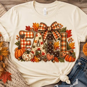 Puede incluir: Una camiseta de color crema con la palabra FALL en letras grandes y decorativas. Las letras tienen estampados de cuadros vichy rojos y blancos, hojas de otoño y lunares marrones. Un lazo de cuadros, calabazas y follaje otoñal rodean el texto.
