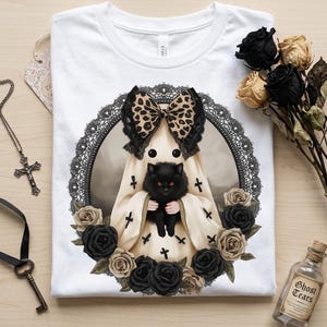 May include: A white t-shirt featuring a graphic of a ghost wearing a leopard print bow and holding a black cat, surrounded by a black lace frame and black and beige roses. The design includes small black cross symbols on the ghost's body.