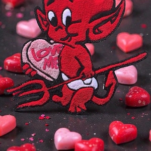 May include: A red devil patch with white horns and a trident, holding a heart-shaped candy that says "Love Me". The patch is surrounded by red and pink heart-shaped candies on a dark surface.