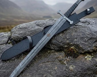 May include: A double-edged sword with a patterned Damascus steel blade, a crossguard, and a wrapped handle, accompanied by a black leather scabbard featuring adjustable straps and a metal stud closure, resting on a dark, textured stone surface.