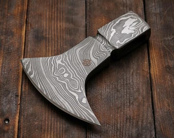 May include: A handcrafted axe with a distinctive pattern on the blade and head. The axe features a curved blade and a wooden handle. The blade has a unique, swirling design in shades of gray and silver. The axe is displayed on a wooden surface.