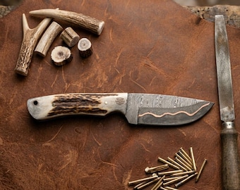 May include: A fixed-blade knife with a textured antler handle and a Damascus steel blade featuring a wavy copper inlay. The knife is accompanied by several antler pieces, a metal file tool, and a small pile of brass pins on a brown surface.
