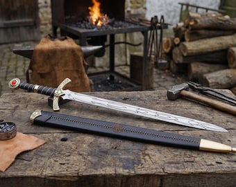 May include: A decorative sword with a black handle, gold accents, and a silver blade with an etched design. The sword rests on a wooden surface next to its black leather sheath. A red gem adorns the pommel.