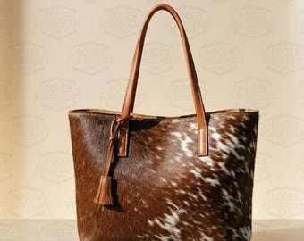 May include: A brown and white cowhide tote bag with brown leather handles and a tassel. The bag has a rectangular shape and a spacious interior, perfect for everyday use. The bag is made of genuine cowhide.