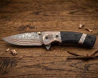 May include: A Damascus steel folding knife with a copper and black handle. The blade has a unique pattern with copper accents. The handle is black with copper and silver details. The knife is on a wooden surface.