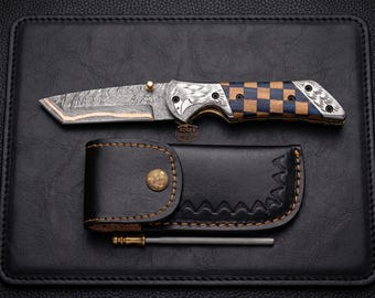 May include: A Damascus steel folding knife with a checkered blue and brown handle, a black leather sheath, and a sharpening rod. The knife blade has a unique pattern. The sheath has a decorative edge and a brass button.