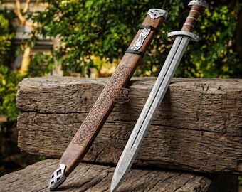 May include: A Viking-style sword with a silver blade and a wooden handle, next to a brown scabbard with Celtic knotwork. The scabbard has silver accents. The sword and scabbard are resting on a weathered wooden surface.