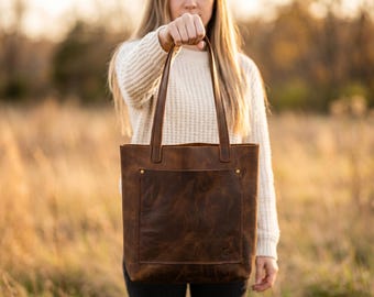 May include: A brown leather tote bag with a front pocket and two handles is held up in front of a blurred background. The bag has a distressed finish and gold-colored rivets. The tote bag is a classic style.