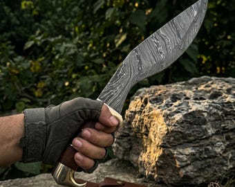 May include: A large, curved knife with a patterned blade is held in a gloved hand. The knife has a wooden handle with brass accents. A brown leather sheath rests below the knife. The background features green foliage and a large rock.