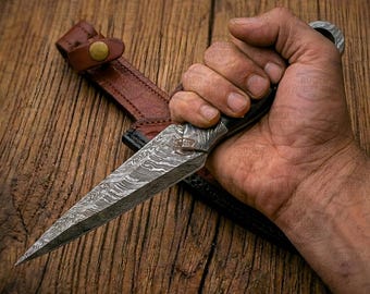May include: A Damascus steel dagger with a patterned blade, held in a hand. The dagger has a black handle and a leather sheath with a brown strap. The background is a wooden surface.