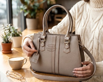 May include: A taupe leather handbag with a top handle and a detachable shoulder strap. The bag features a rectangular shape with vertical stitching details and the text "ROGER FORGEKRAFT". A small tag hangs from the handle.