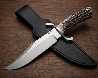 May include: A large, stainless steel hunting knife with a bone handle and a black leather sheath. The knife has a curved blade and a guard. The sheath is stitched and has a belt loop. The knife and sheath are on a wooden surface.