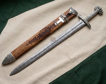 May include: A detailed overhead shot of a Viking sword and scabbard. The sword has a silver handle and a patterned blade. The scabbard is made of wood with metal accents and intricate carvings. The overall color scheme is brown, silver, and gray.