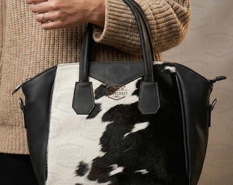 May include: A black and white cowhide handbag with black leather accents. The bag has two handles and a shoulder strap. The brand name "ROGER FORGECRAFT" is visible on the front. The bag is held by a person wearing a tan sweater.