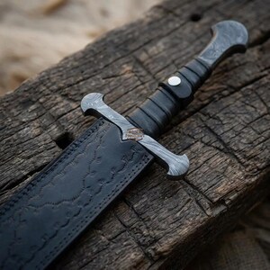 May include: A fixed-blade knife featuring a patterned Damascus steel crossguard and pommel, a black wrapped handle with a silver rivet, and a black leather sheath embossed with a repeating pattern. The blade bears the text Roder Forgecraft.