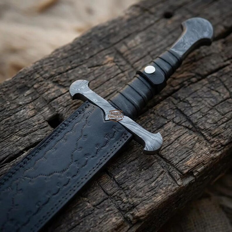 May include: A fixed-blade knife featuring a patterned Damascus steel crossguard and pommel, a black wrapped handle with a silver rivet, and a black leather sheath embossed with a repeating pattern. The blade bears the text Roder Forgecraft.