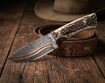 May include: A handcrafted hunting knife with a patterned blade and a handle made of antler material. The blade has a copper-colored stripe. The knife rests on a brown leather belt, with a cowboy hat in the background.