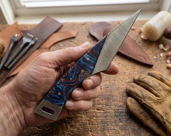 May include: A handcrafted knife with a stainless steel blade and a handle featuring a blue and orange swirl pattern. The knife is held in a hand, with leatherworking tools and a mallet in the background.