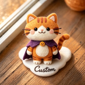 Könnte beinhalten: Eine nadelgefilzte orangefarbene getigerte Katzenfigur, die einen violetten Umhang mit einem kleinen orangefarbenen Kürbisanhänger am Hals trägt. Die Katze steht auf einer weißen wolkenförmigen Basis mit dem Wort Custom in schwarzem Garn.