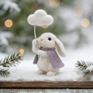 Może przedstawiać: Biała filcowa figurka królika w liliowej pelerynie, trzymająca białą chmurę na sznurku. Scena jest zaśnieżona, z gałązkami sosny i światłami bokeh w tle. Fantazyjny, ręcznie robiony przedmiot.