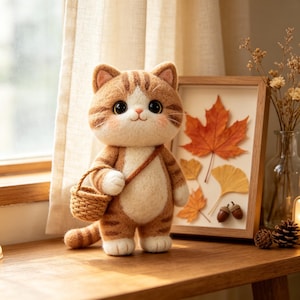 May include: A needle-felted orange tabby cat figurine with white paws and chest, featuring large black eyes and a small pink nose. The cat carries a miniature woven basket on its arm and has a striped tail, standing upright with a soft, fuzzy texture.