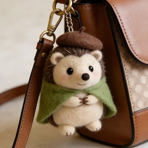 May include: A handmade hedgehog keychain with a brown beret and a green cape. The hedgehog has a cream-colored body, brown quills, and rosy cheeks. It is attached to a gold-colored chain and clasp, hanging next to a brown leather purse.