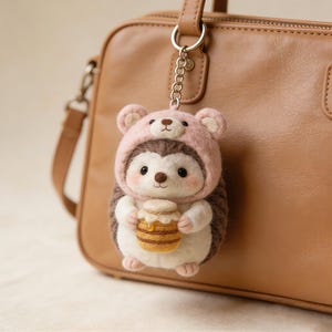 Könnte beinhalten: Ein handgefertigter Igel-Schlüsselanhänger mit einem rosa Bärenhut, der einen Honigtopf hält. Der Schlüsselanhänger ist an einer braunen Handtasche befestigt. Der Igel hat einen braunen und weißen Körper mit schwarzen Augen und einem lächelnden Gesicht.