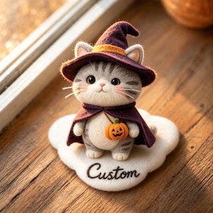 Könnte beinhalten: Eine nadelgefilzte graue getigerte Katzenfigur mit einem violetten Hexenhut mit orangefarbenem Band und einem passenden violetten Umhang. Die Katze hält einen kleinen orangefarbenen Kürbis. Sie steht auf einer weißen Filzbasis mit dem Wort Custom.