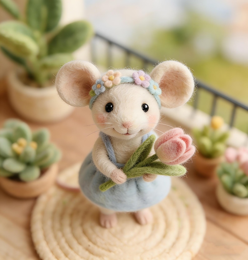 May include: A handcrafted white mouse figurine wearing a light blue dress and a floral headband. The mouse is holding a pink tulip and is standing on a woven mat. The background includes potted plants and a balcony railing.