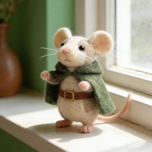 May include: A handcrafted felt mouse figurine with a white and tan body, wearing a green cape and a brown belt with a gold buckle. The mouse has large ears, a pink nose, and a long pink tail. The figurine is standing on a white surface.