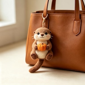 Könnte beinhalten: Ein nadelgefilzter Otter-Schlüsselanhänger mit hellbraunem und cremefarbenem Körper, der einen kleinen orangefarbenen Kürbis hält. Der Otter hat dunkle Augen, eine gestickte Nase und Schnurrhaare und ist an einem goldfarbenen Metallclip befestigt.